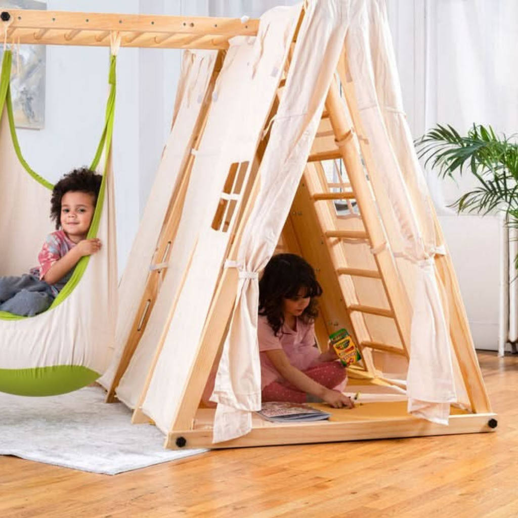 Children playing inside a wooden teepee with a hammock in a room.