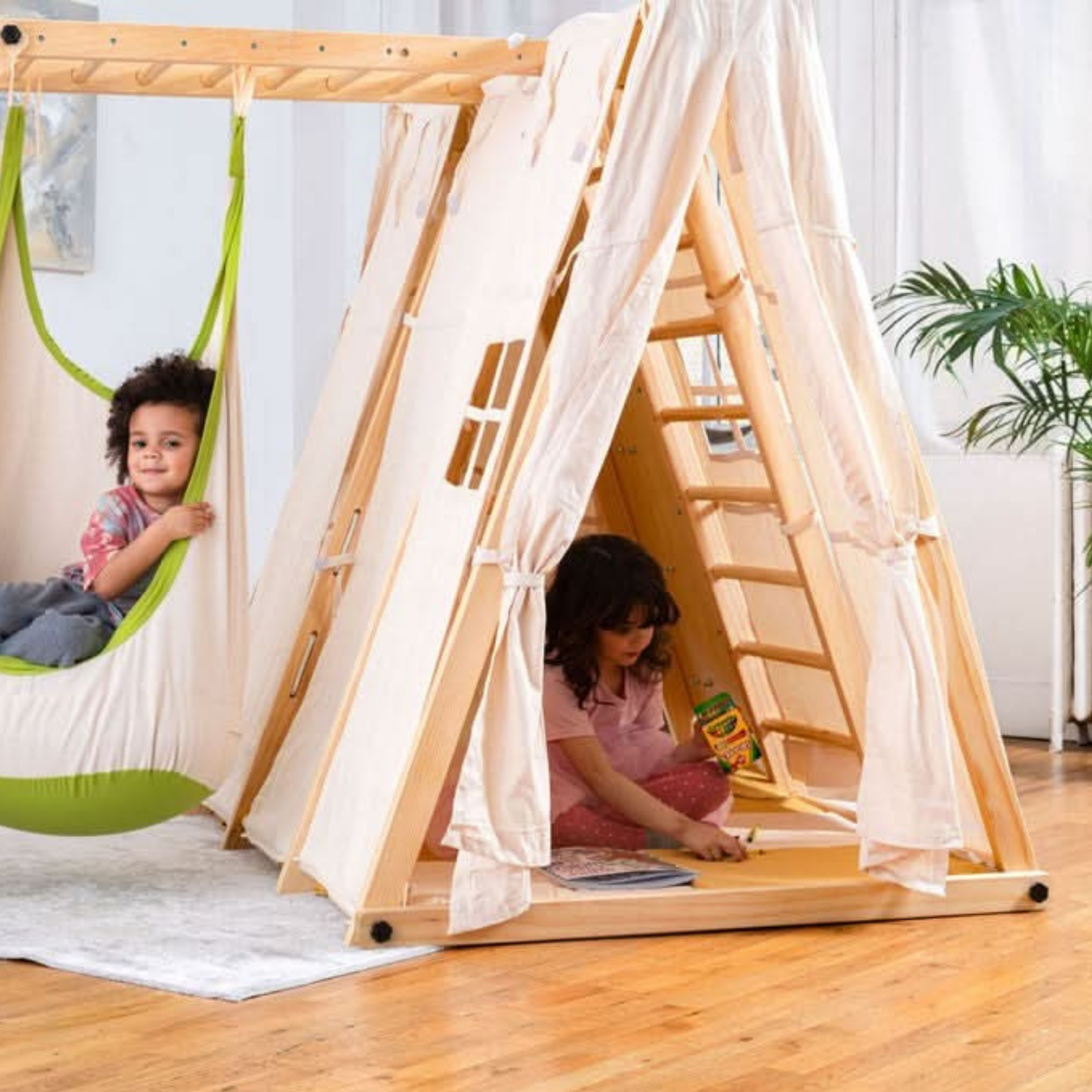 Children playing inside a wooden teepee with a hammock in a room.