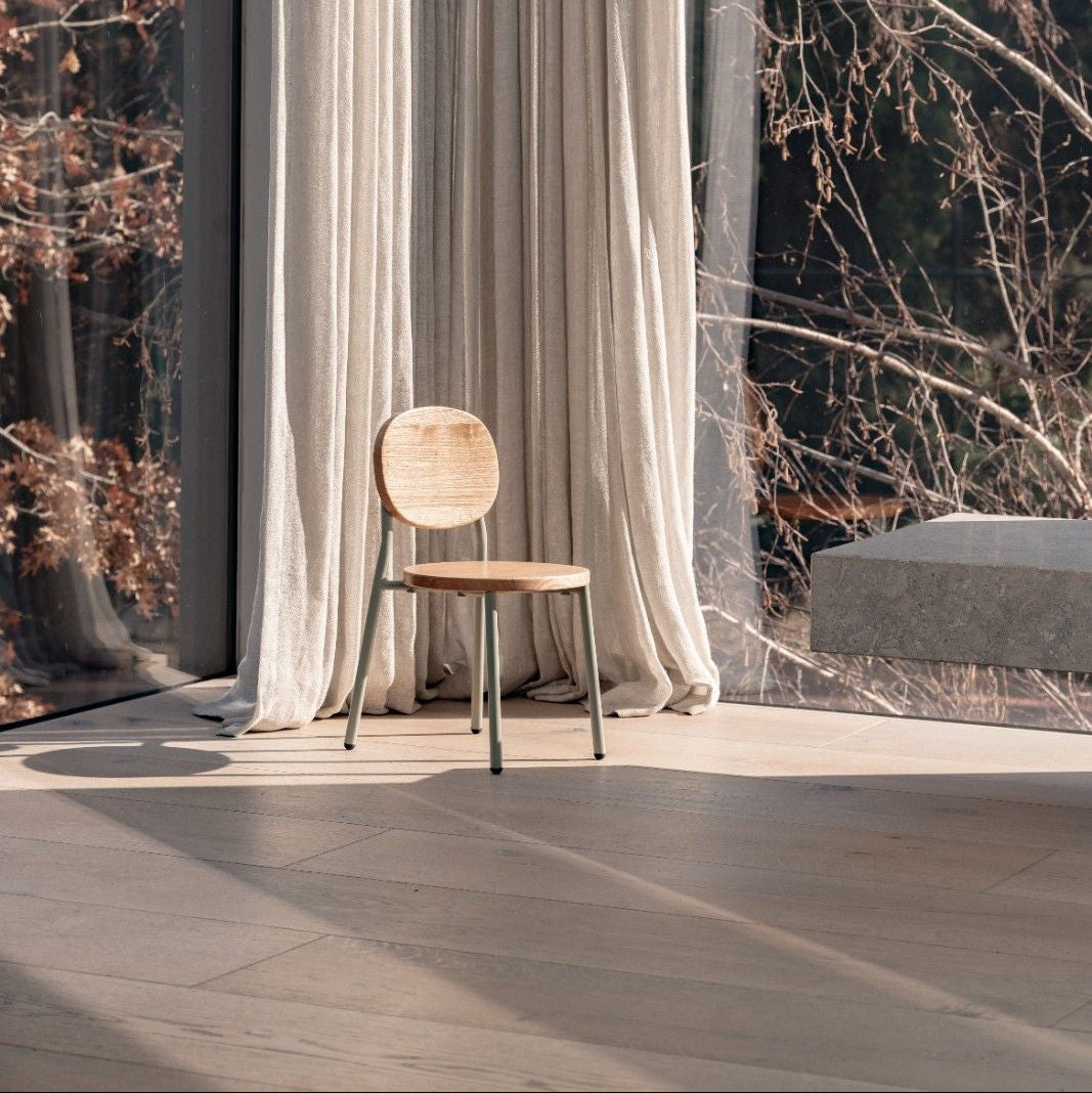 Wooden chair in front of a large window with white curtains and a view of trees.