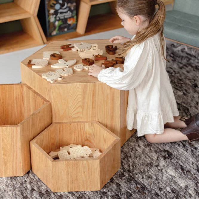 Child playing with wooden toys on a hexagonal wooden table in a room with shelves and books.