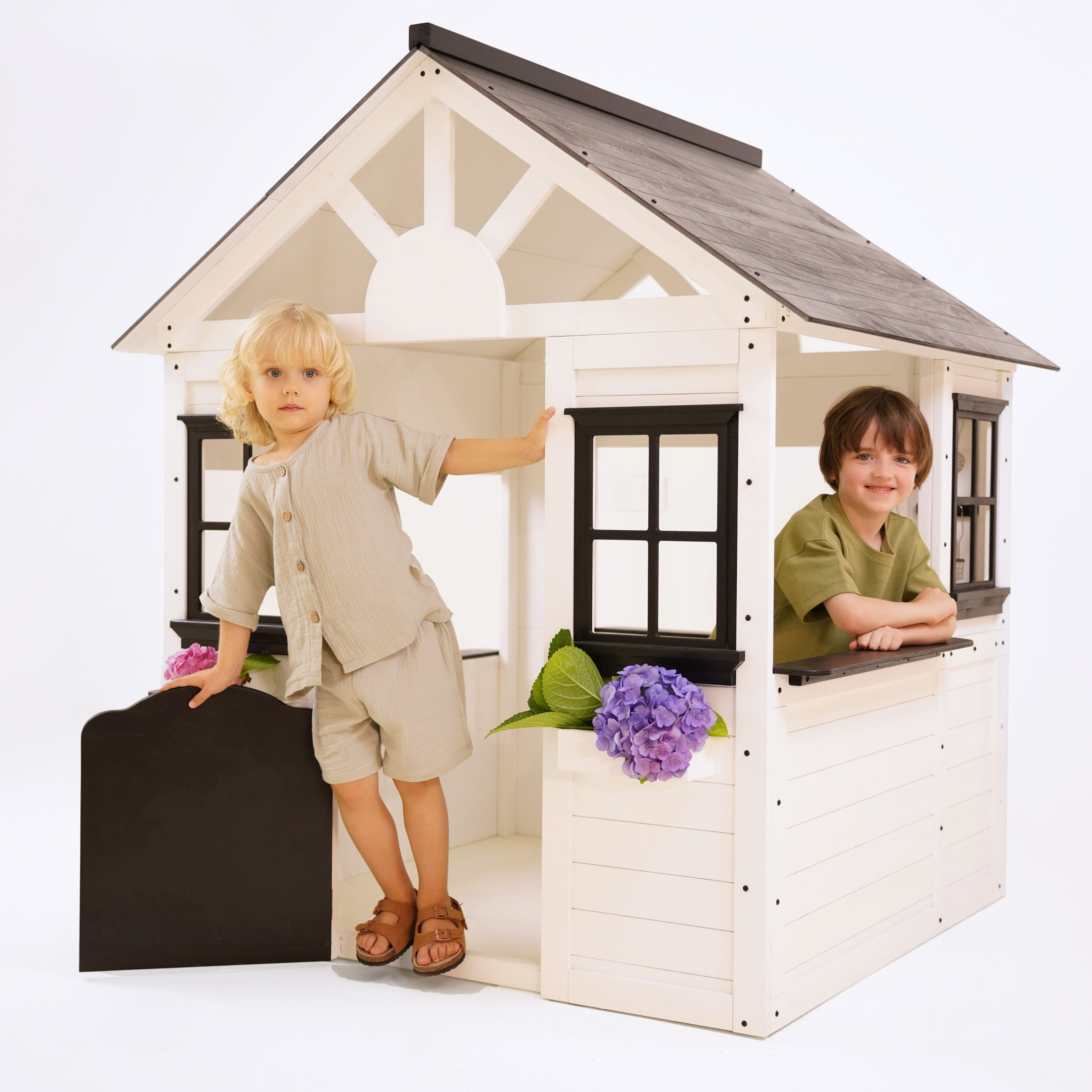 Blonde child standing at the doorway and another child leaning out the side window of a white wooden playhouse decorated with purple and pink flowers