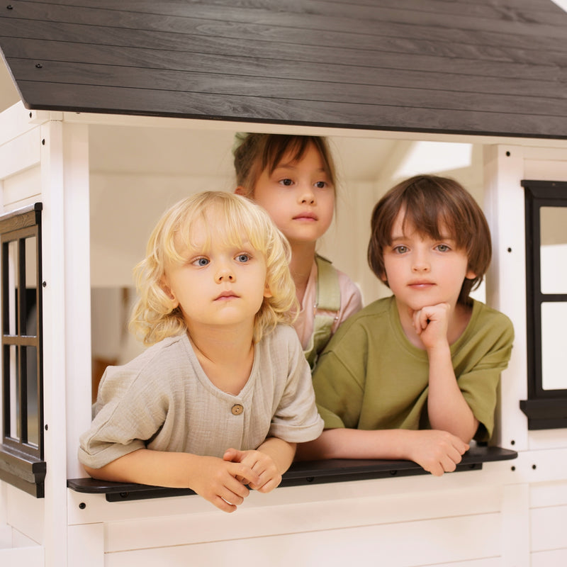 Three children leaning out of the window of a white wooden playhouse with black trim, smiling and looking outward.