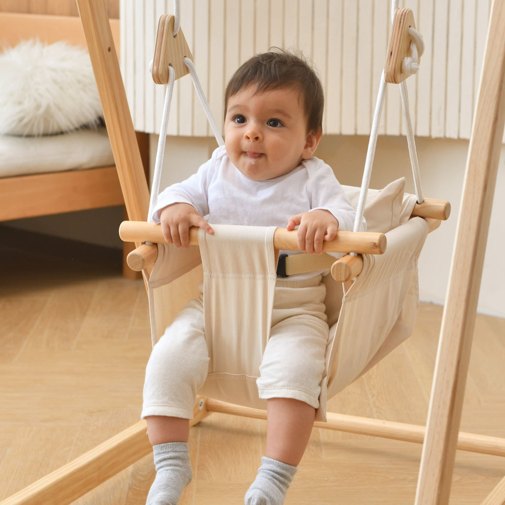 Baby sitting in the Avenlur off white cotton canvas hammock swing seat with wooden front bar, hanging from a wooden indoor swing stand
